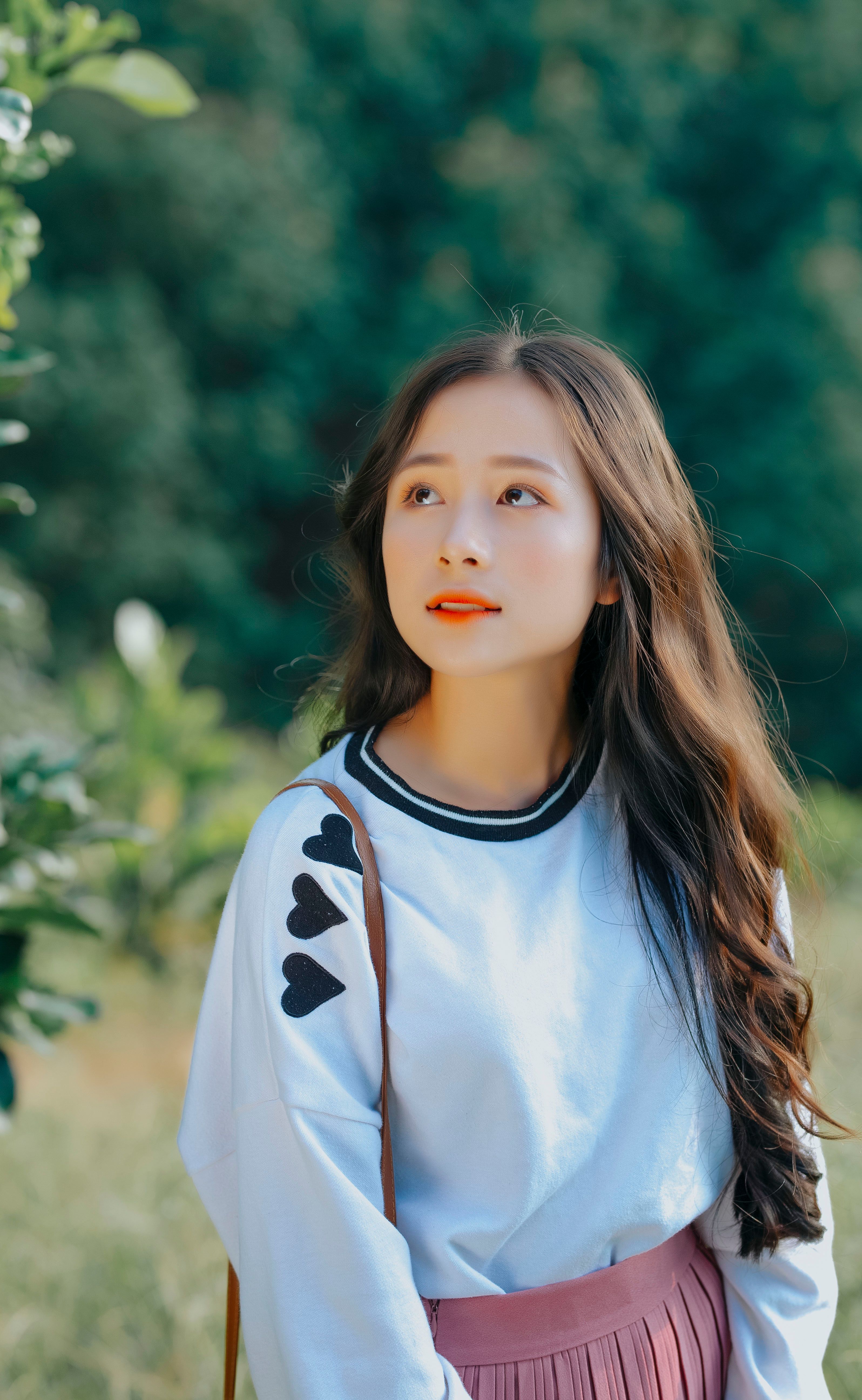Asian woman with long hair wearing a white top with heart detail, looking pensive outdoors.