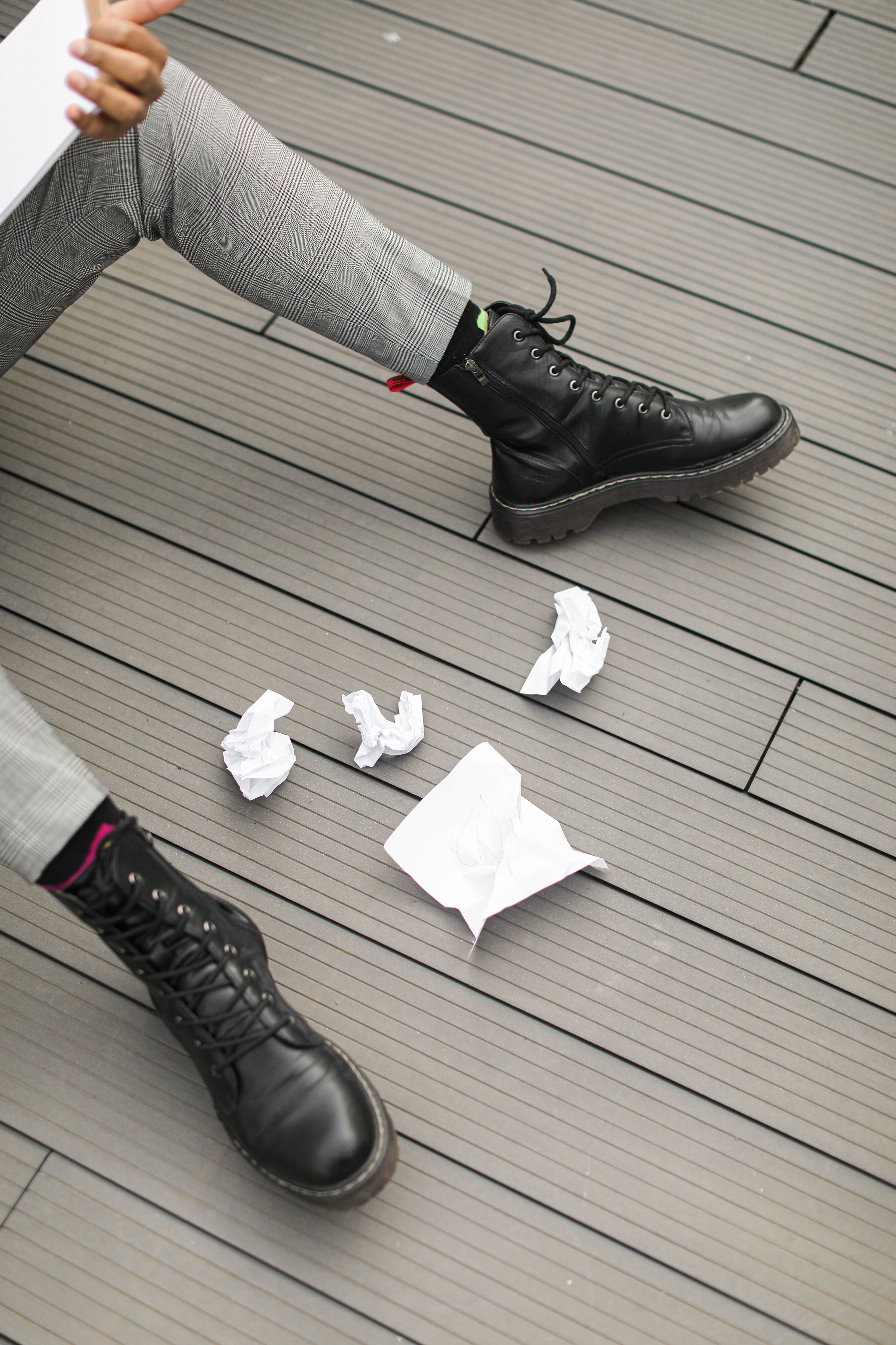 Black boots and legs with crumpled papers on a wooden floor indoors.