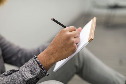 An adult hand writes in a notebook with a pencil, wearing a beaded bracelet and gray plaid sleeves.