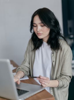 Young woman using credit card for online shopping at home, sitting at a wooden table with laptop.