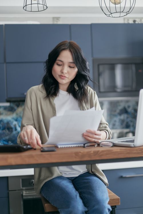 Free A young woman reviews documents and uses her laptop in a modern kitchen setting. Stock Photo