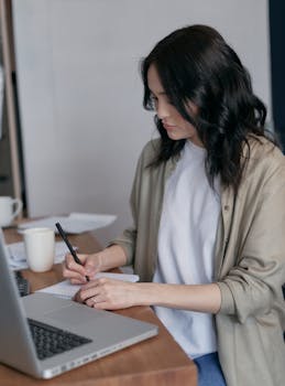 Focused woman taking notes at a laptop in an office setting, emphasizing productivity.