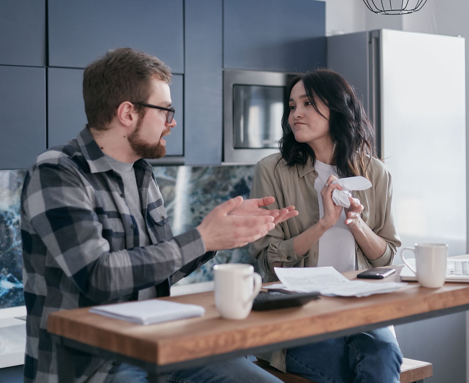 Homem e mulher sentados à mesa da cozinha olhando comprovantes de compras e digitando no celular.