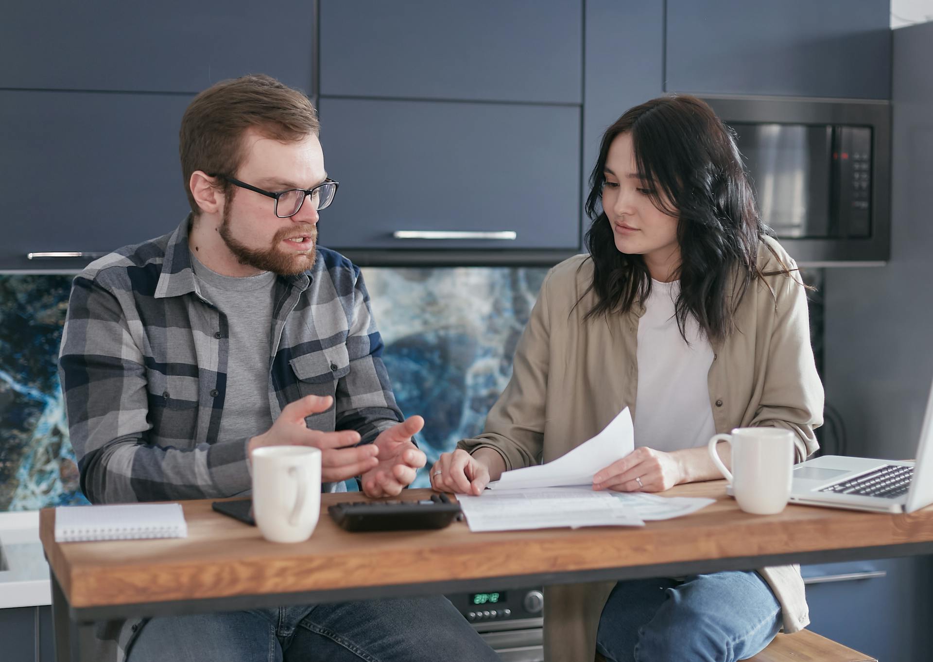 A couple engaged in a financial discussion, sitting at a kitchen table with papers and a laptop.