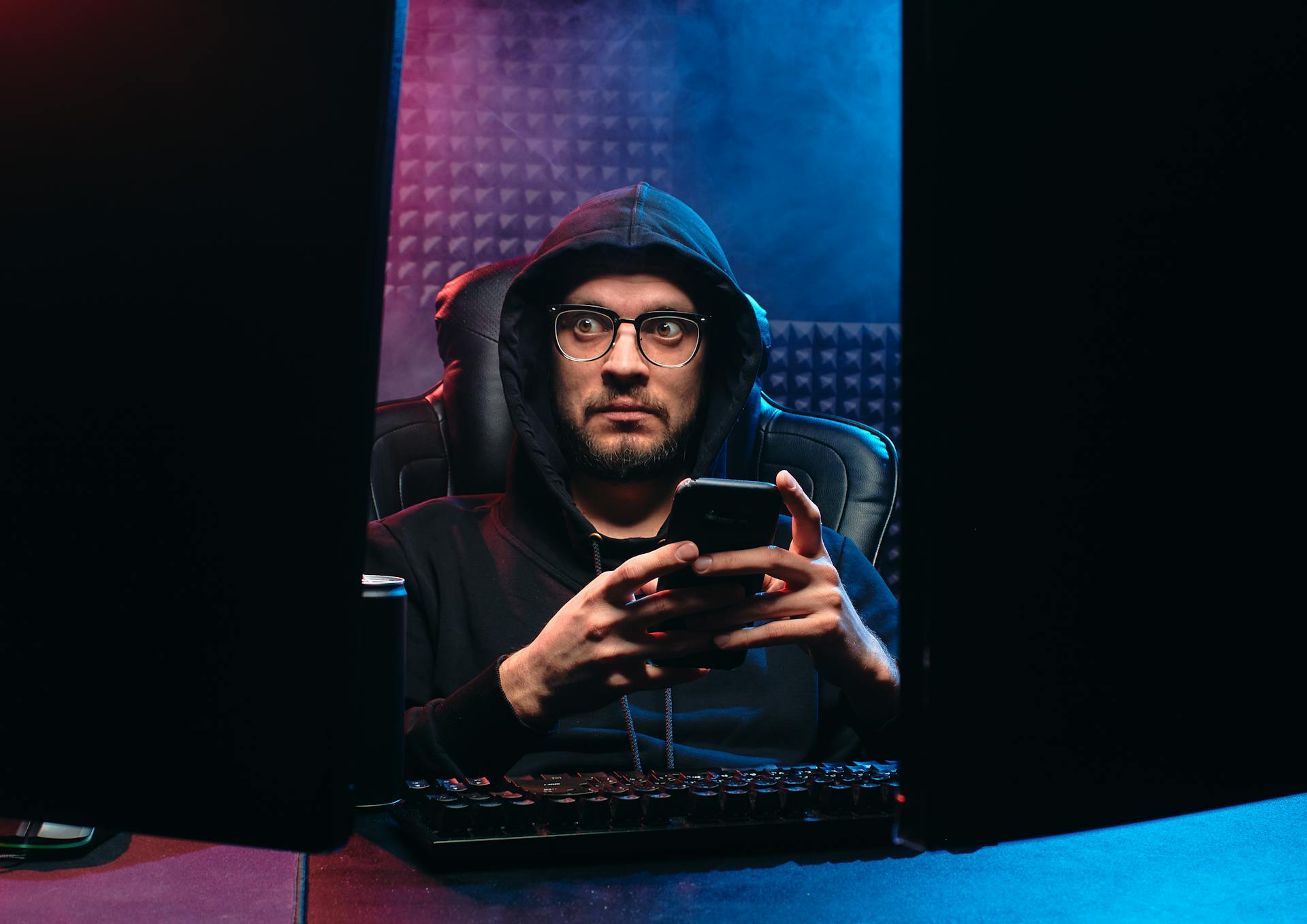 Man in hoodie with eyeglasses using smartphone between computer screens in a dark room.