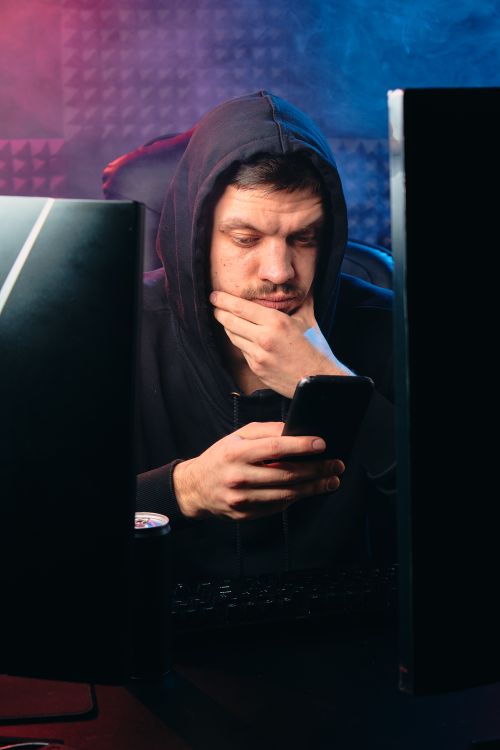 Free Concentrated man in hoodie looking at smartphone between two monitors. Stock Photo