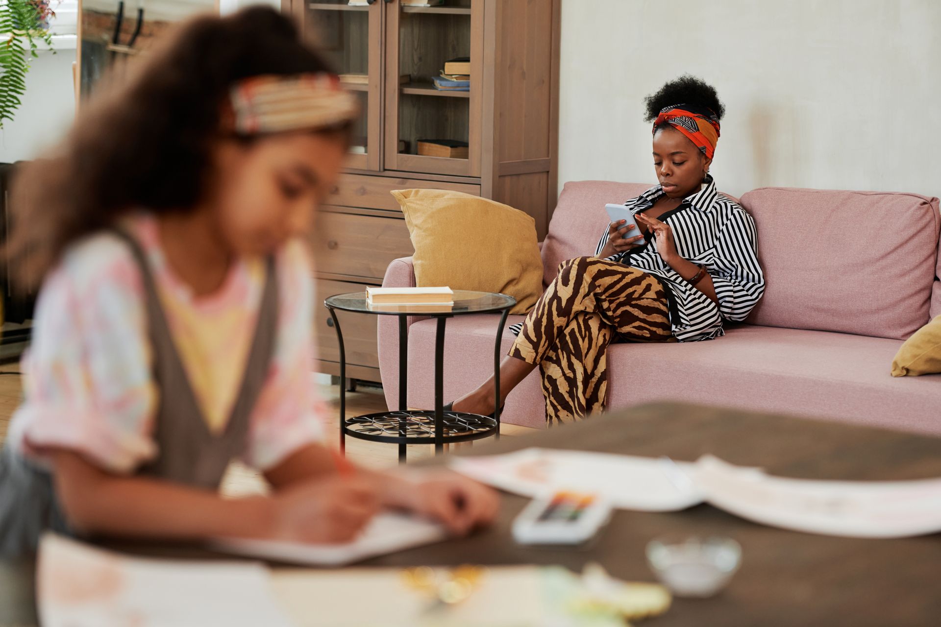 Mother texting on sofa, child drawing at table, in cozy home setting.