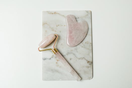 Flat lay of rose quartz facial roller and gua sha stone on marble background for skincare routines.