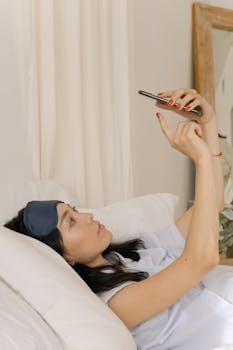 Asian woman lying in bed, using smartphone, wearing sleep mask, showing a peaceful morning routine.