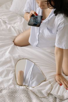Asian woman in pajamas uses a smartphone for a selfie sitting on a bed.
