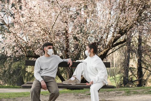Free Young couple wearing face masks sitting on a park bench, practicing social distancing during the pandemic. Stock Photo