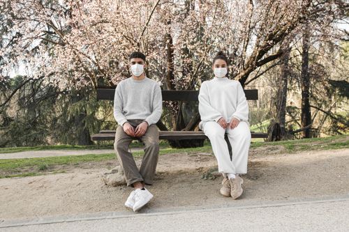 Free A man and woman in masks sit apart on a bench under blooming trees, reflecting social distancing. Stock Photo