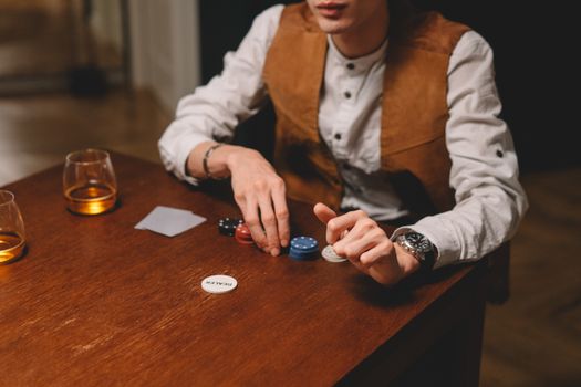 A man playing poker indoors, with poker chips and whiskey on a wooden table.