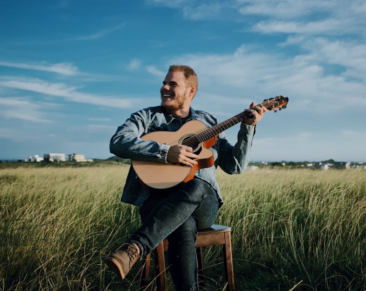 Happy Young Man Playing Guitar In Rural Meadow On Sunny Day