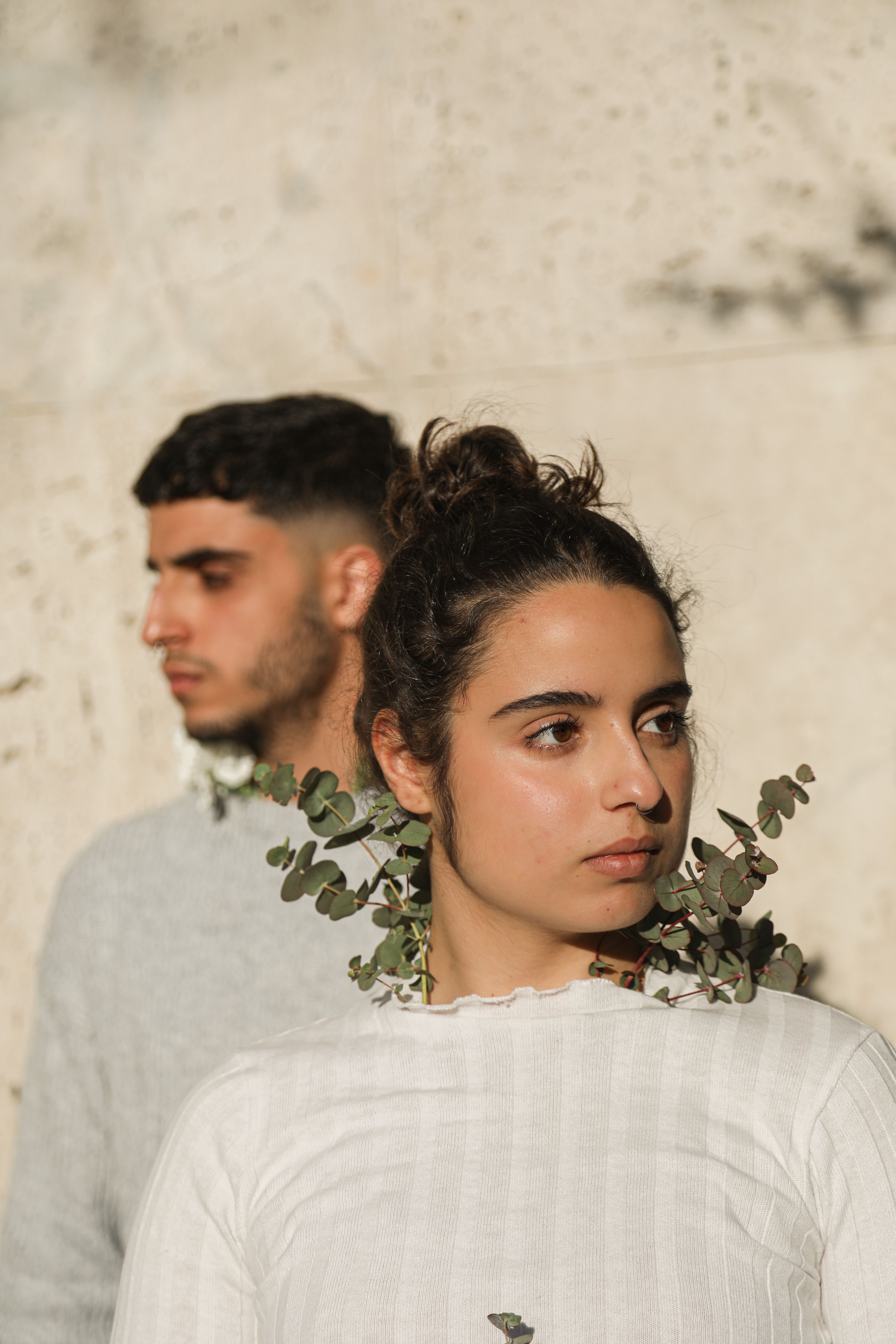 A man and woman standing together, adorned with green leaves, against a concrete wall in natural light.
