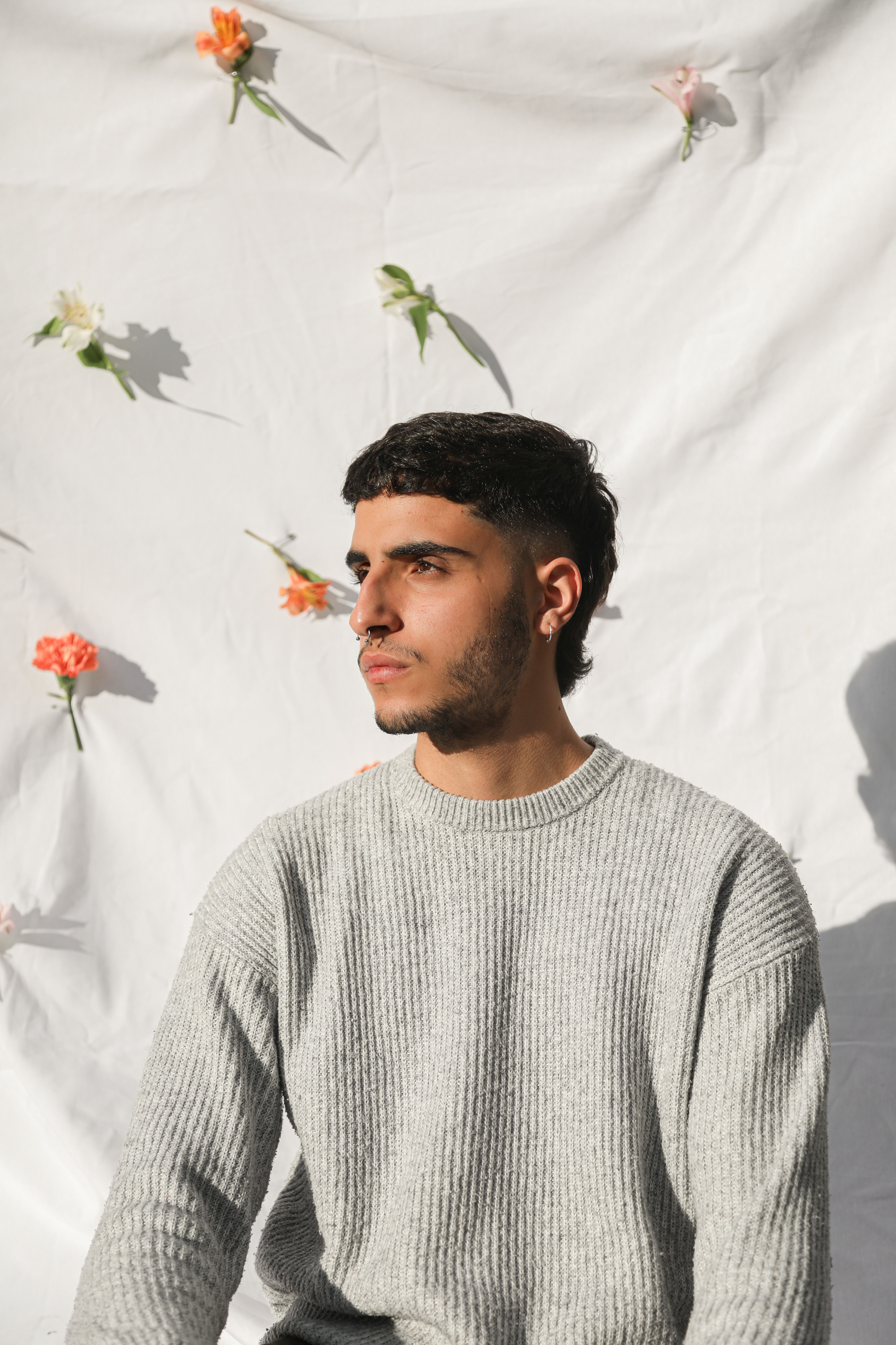 A stylish young man wearing a gray sweater against a white backdrop with colorful flowers.