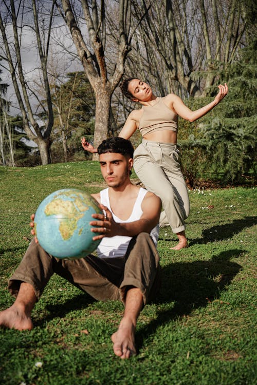 Man Sitting on Grass and Holding a Globe While Girl Dancing Behind