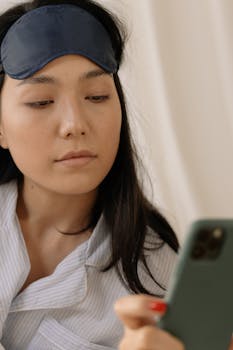 Asian woman in sleepwear uses a mobile phone while wearing a sleep mask indoors.