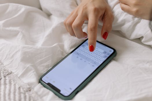 Close-up of woman with red nails typing on a smartphone in bed. Cozy and relaxed setting.