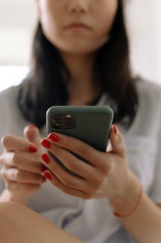 Close-up of a woman holding a smartphone indoors, showcasing red nail polish and a relaxed casual setting.