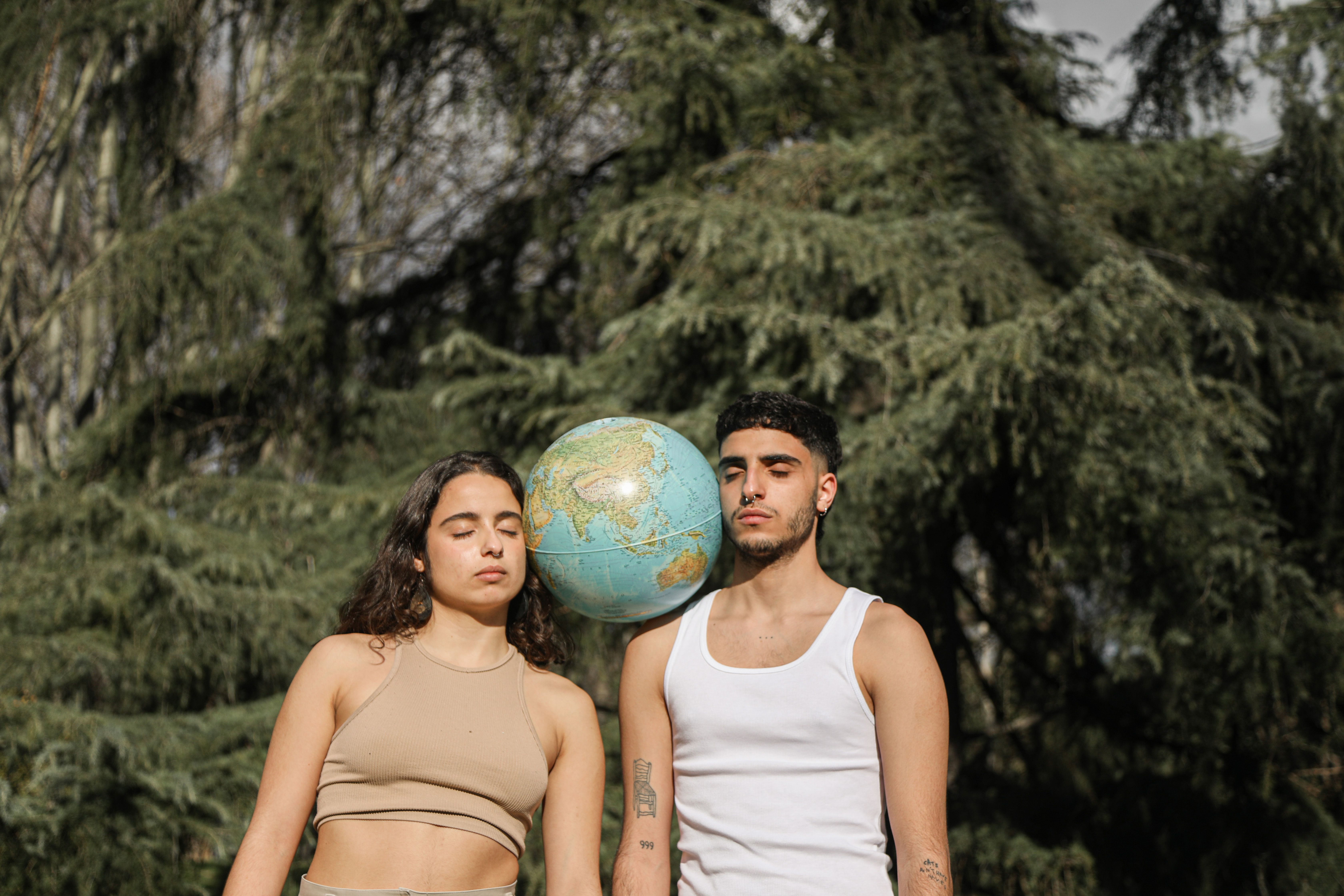 Two young adults standing with a globe amidst trees, eyes closed.
