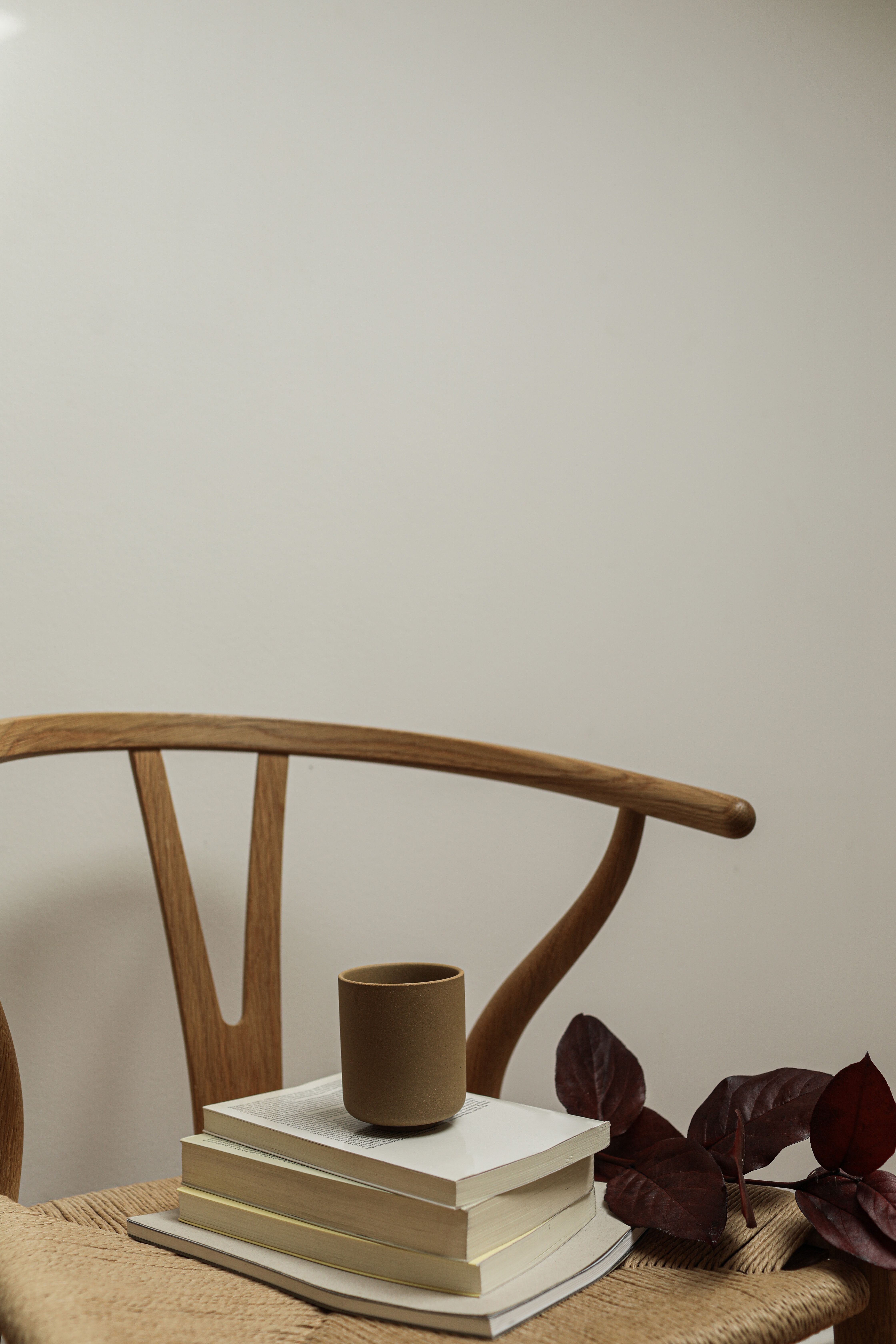 A minimalist setup with a ceramic cup on books beside a wooden chair against a white wall.