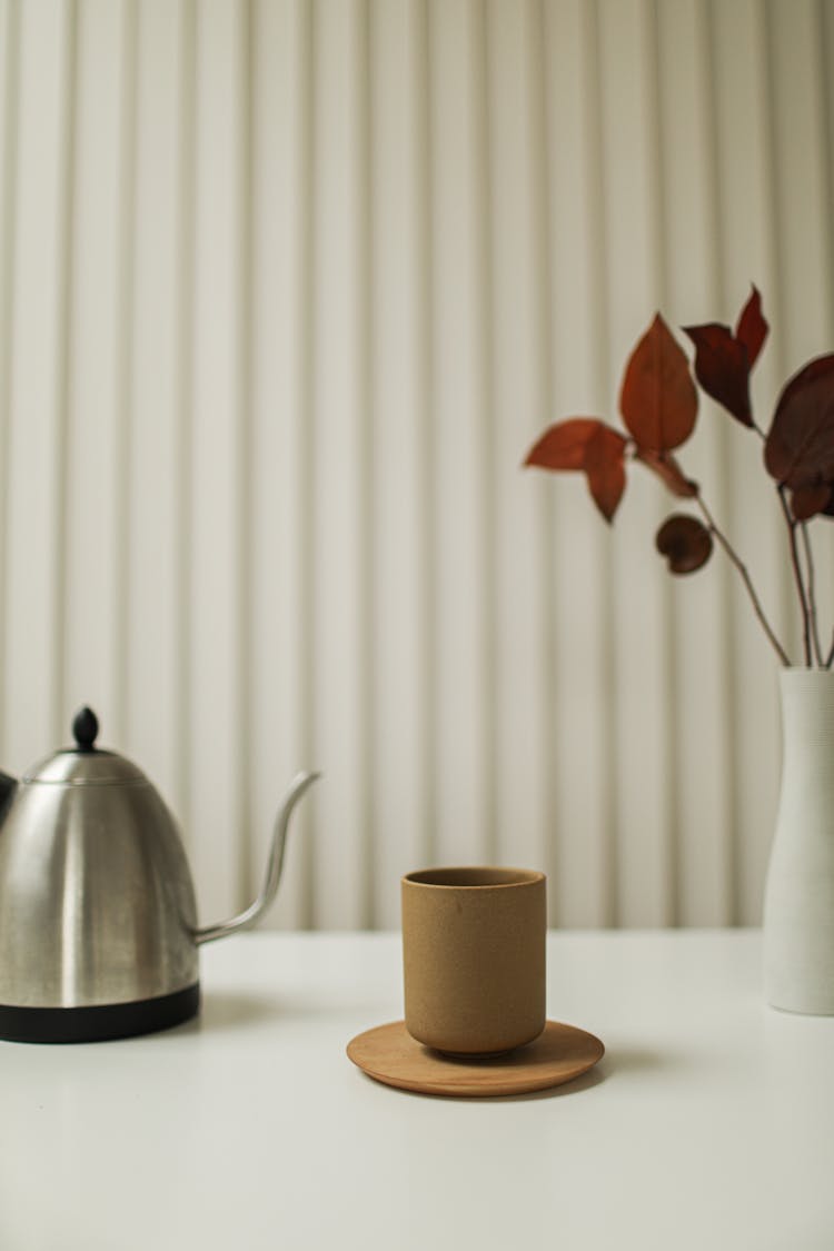 Brown Cup On Brown Wooden Coaster Beside Silver Kettle And White Vase With Red Brown Leaves