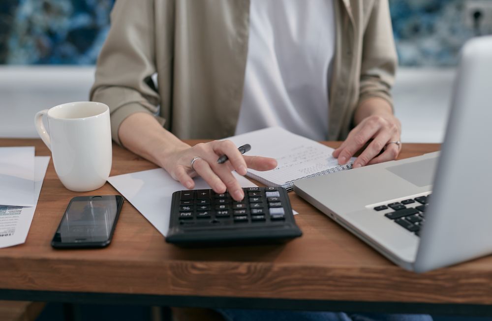 Hands using a calculator and notebook next to a laptop