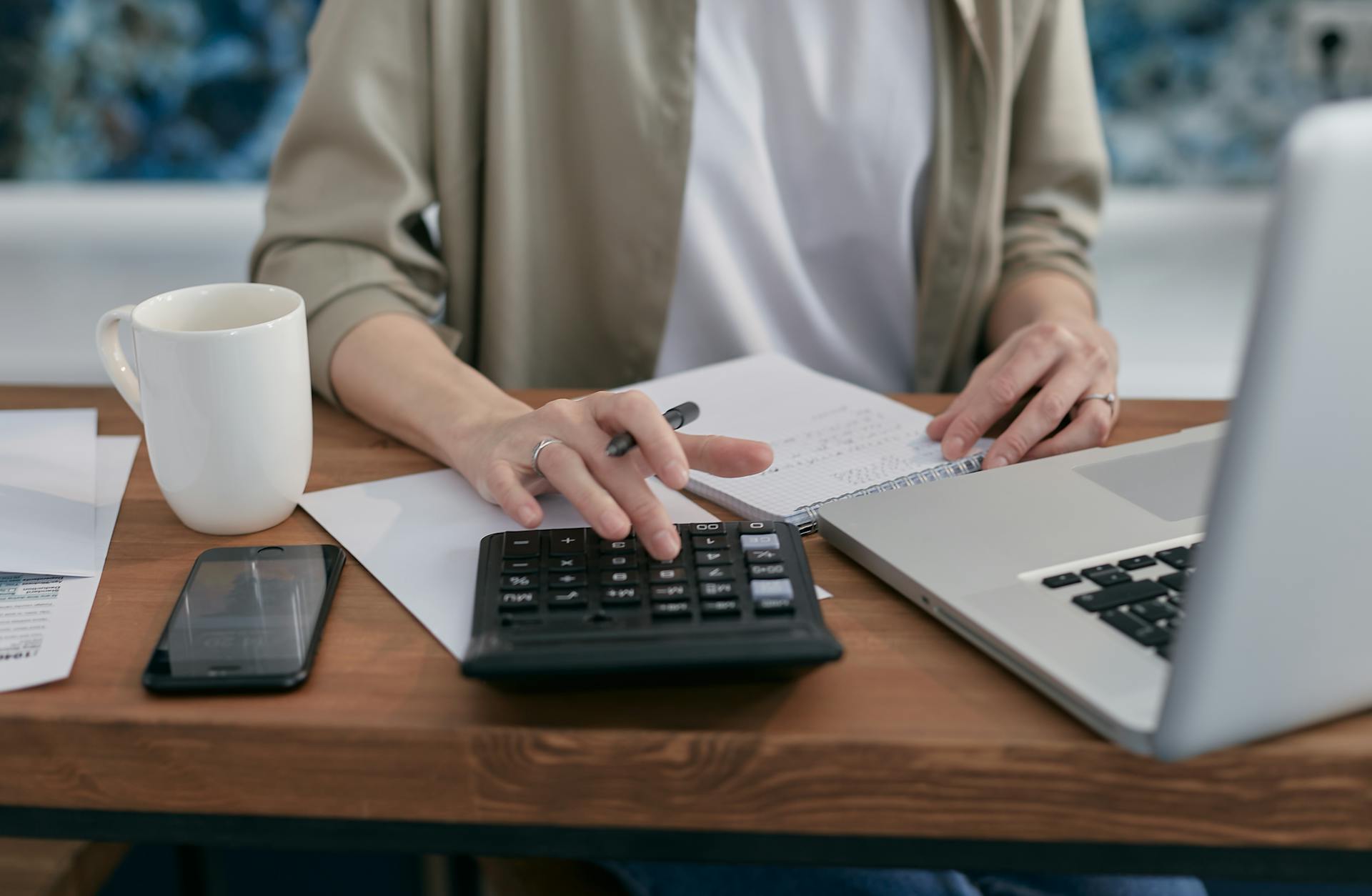 A person using a calculator and laptop while taking notes at a wooden desk.