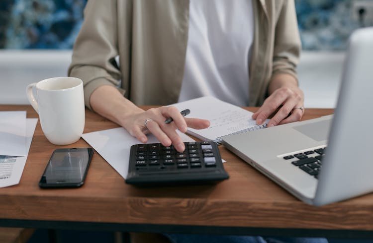 A Person Sitting A Wooden Table With A Notepad And Laptop Using A Calculator