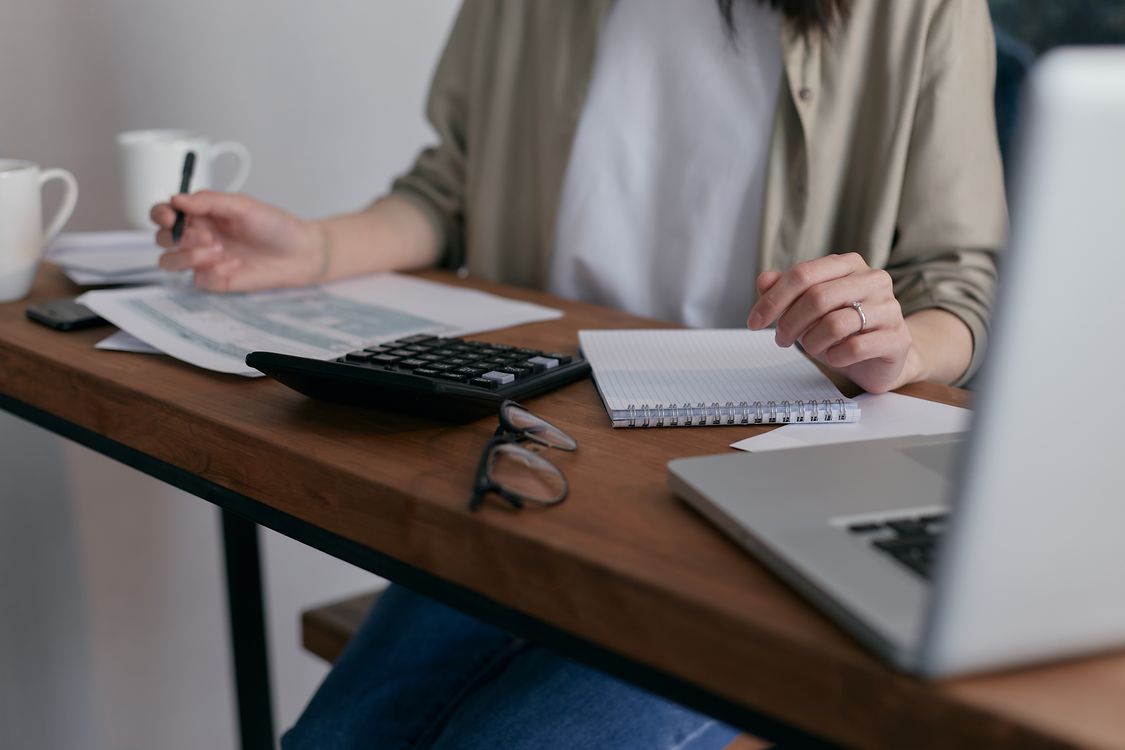 Mulher sentada à mesa fazendo anotações e cálculos, com papéis, calculadora e computador.