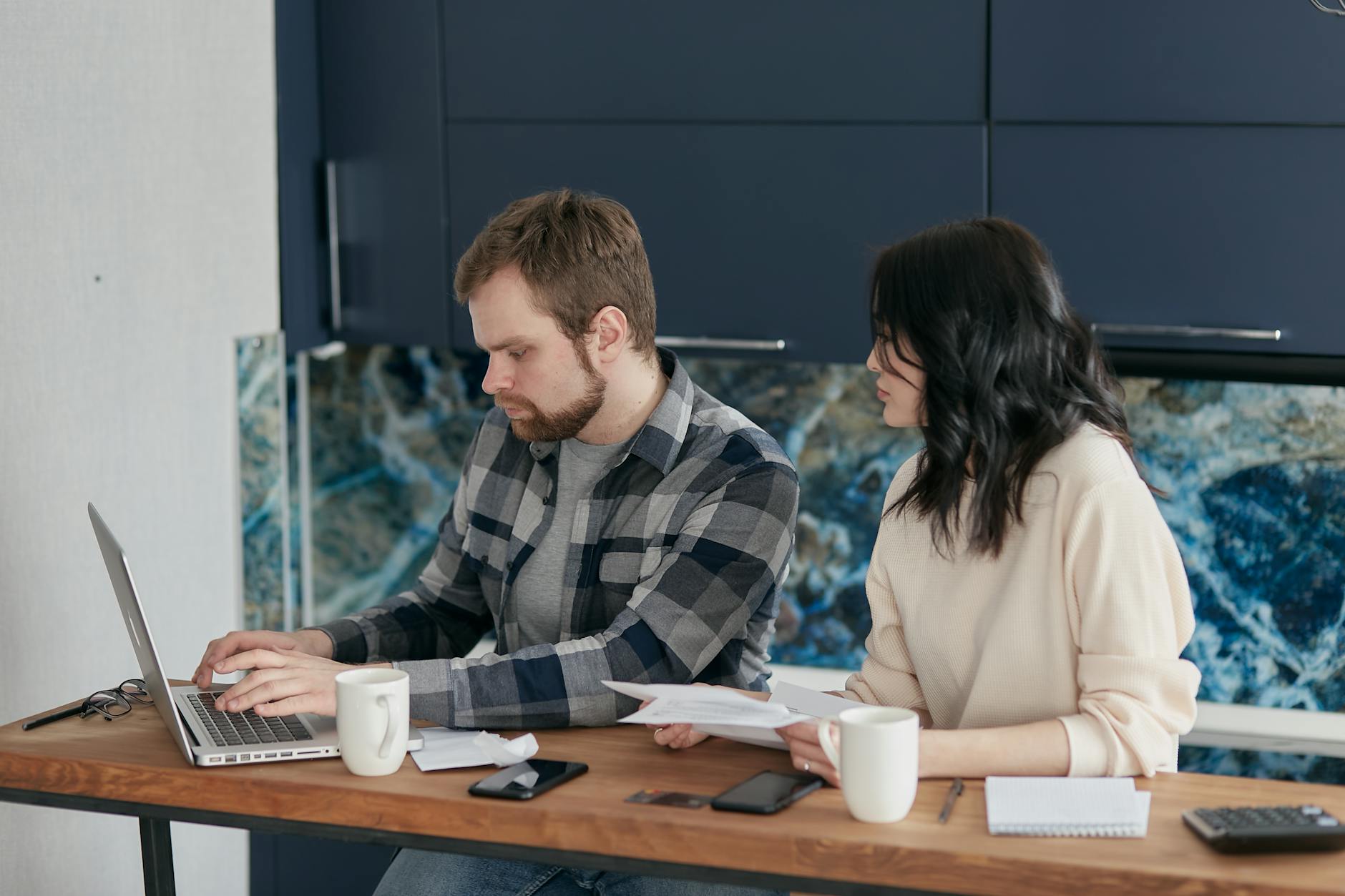 A couple reviews bills and finances at a desk with a laptop and papers. Indoor setting, focused on budgeting.