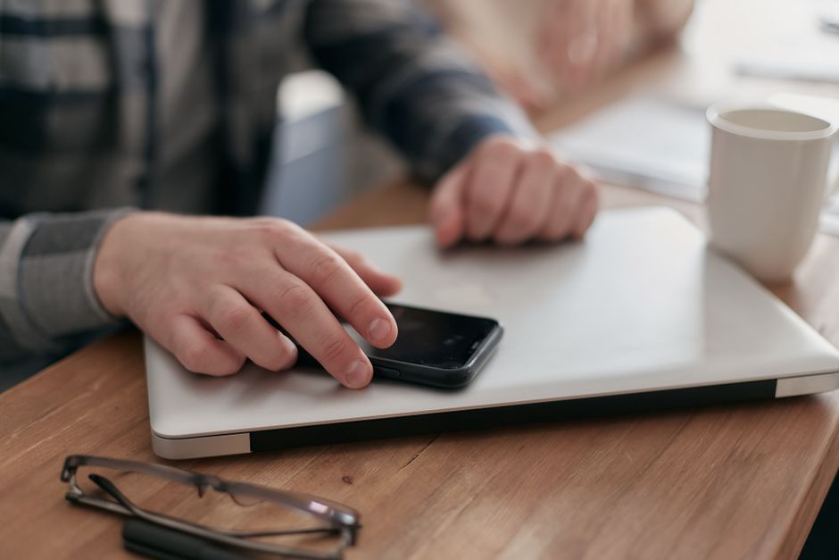 Close-up of hands using smartphone and laptop at a wooden desk with glasses and coffee.