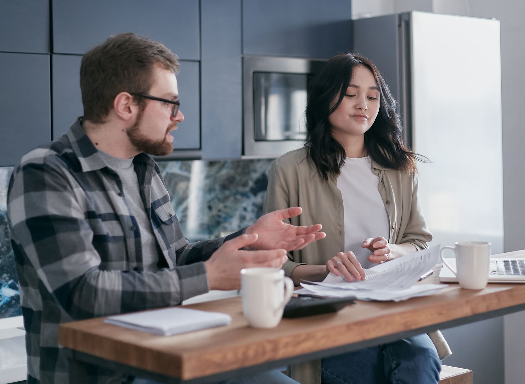 Casal sentado à mesa da cozinha conversando com papéis e celular sobre a mesa.