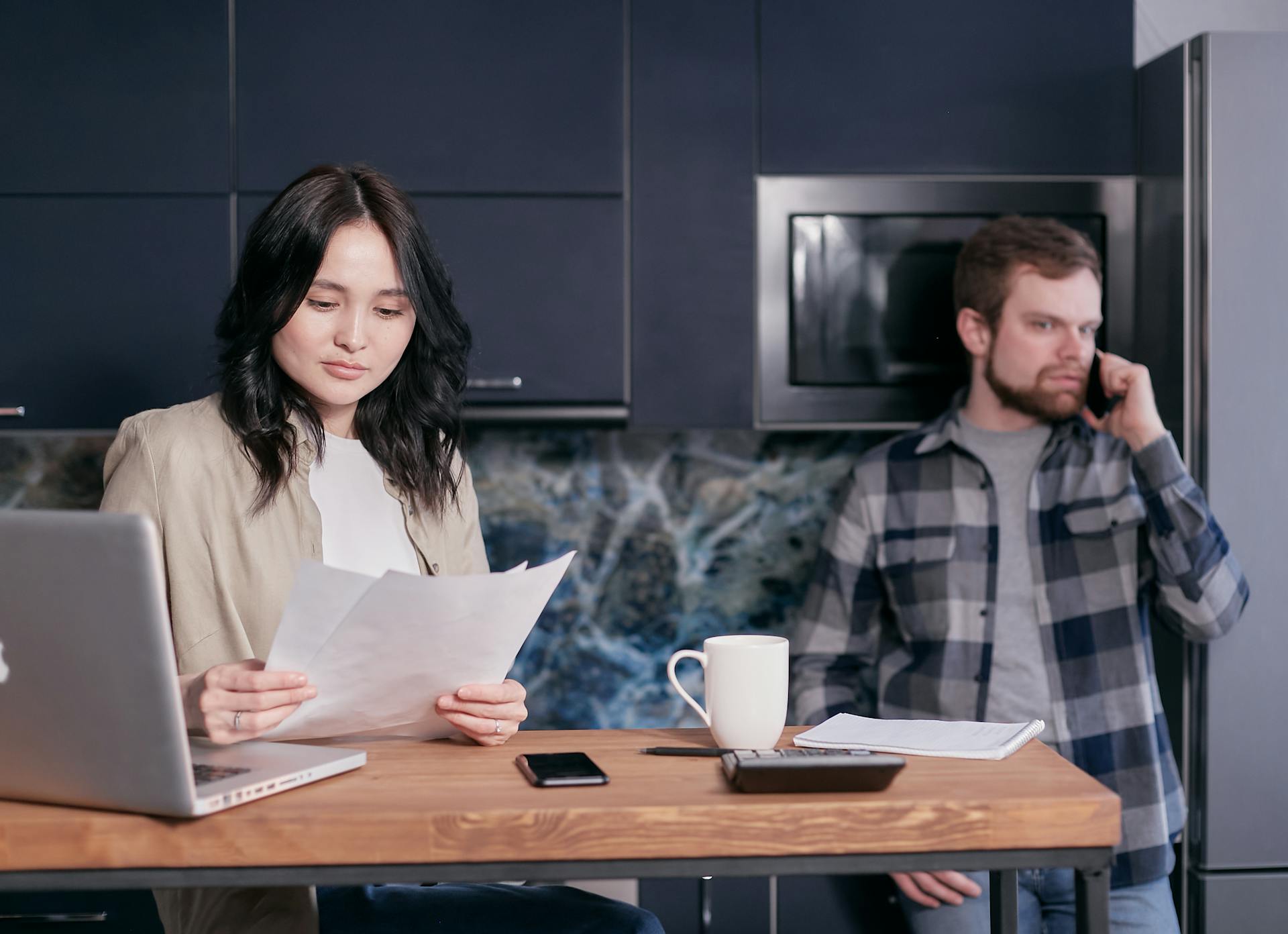 Asian woman and Caucasian man reviewing bills and discussing finances in a modern kitchen setting.