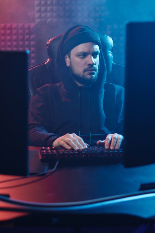Free A focused man in a black hoodie typing on a computer keyboard in a dim, atmospheric room. Stock Photo