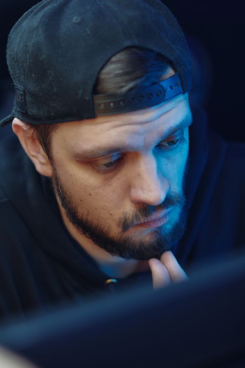 Free A thoughtful man with facial hair wearing a cap illuminated by blue ambient light. Stock Photo