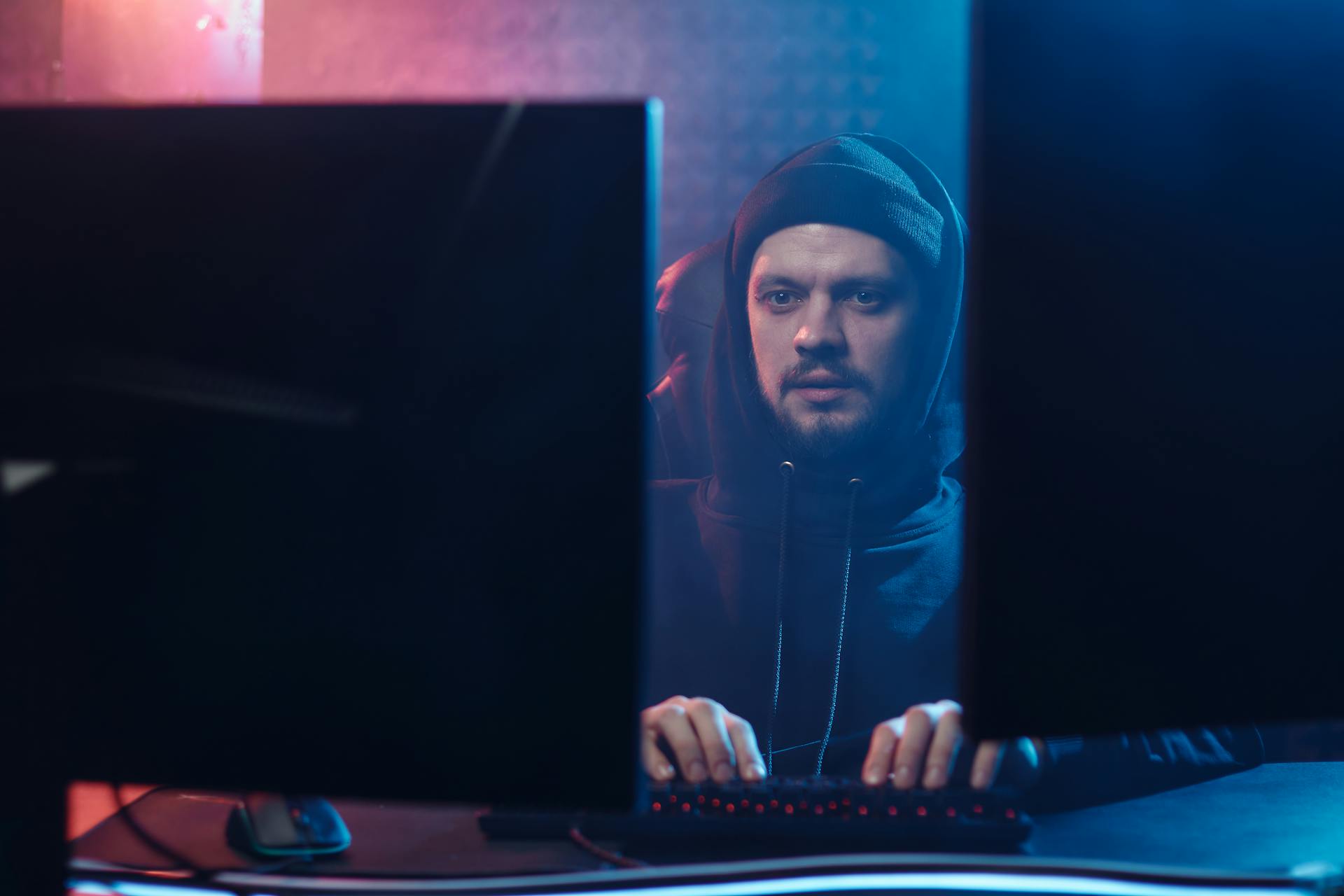 A man with a hoodie typing on a keyboard between two monitors in a dimly lit room.