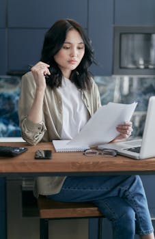 Asian woman in casual attire reading documents at a modern home office desk.