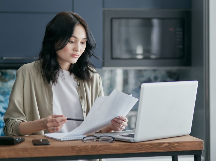 Concentrated woman reviewing documents and working on a laptop at home office setup in kitchen.