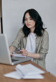 A young adult woman attentively working on her laptop at home, taking notes.