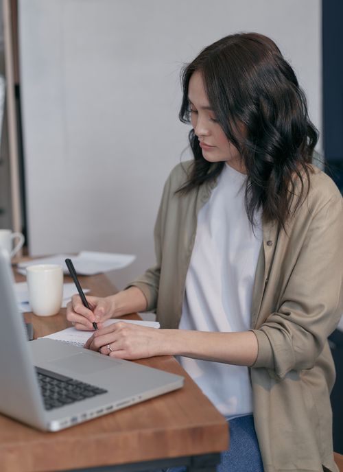Free Adult woman writing notes while using a laptop at her desk in a cozy home office setting. Stock Photo
