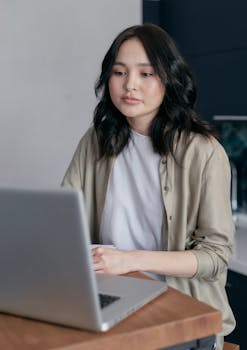 A young woman with black hair working on a laptop at a desk indoors.