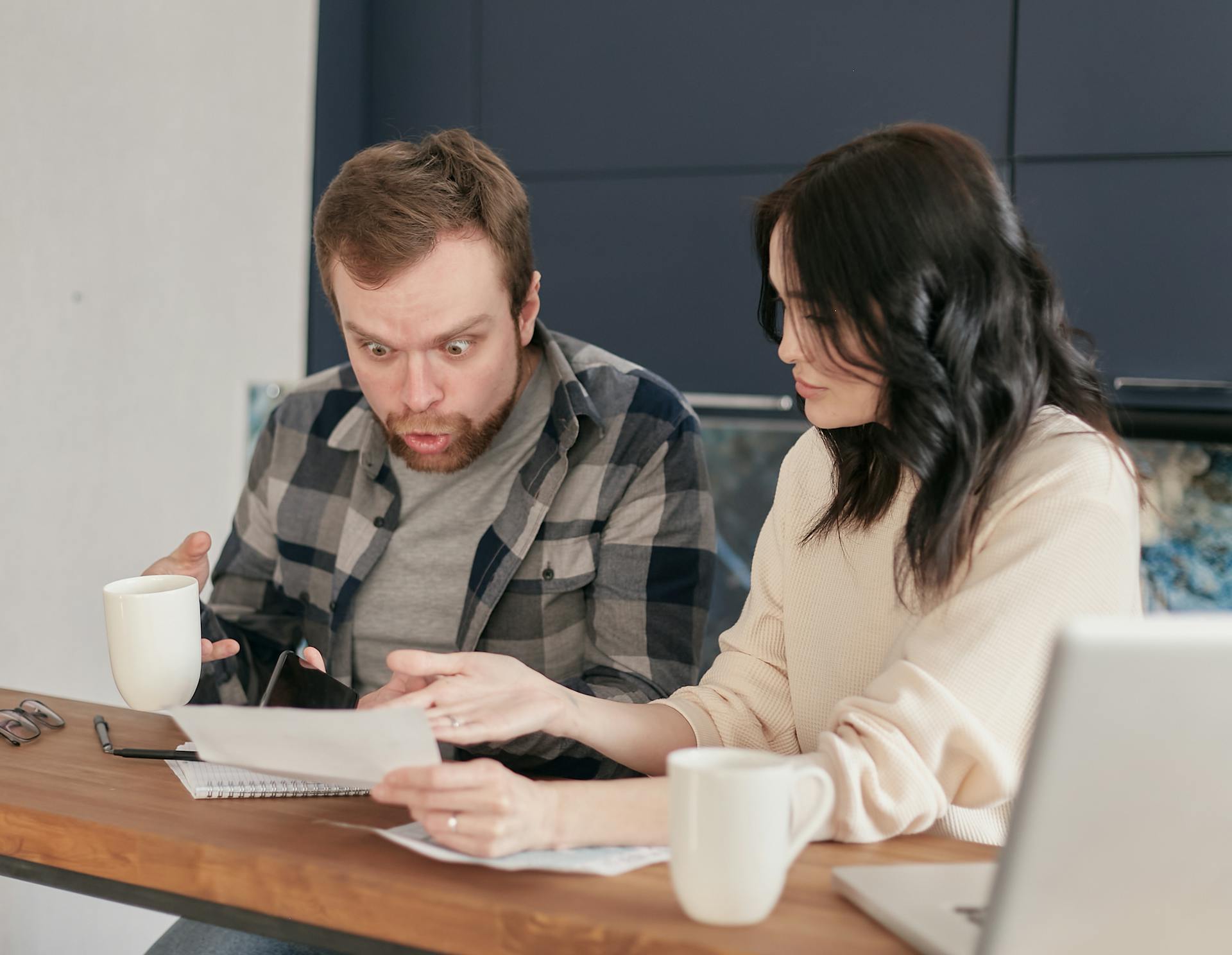 A couple looks shocked while reviewing a document over coffee at home, with a laptop on the table.