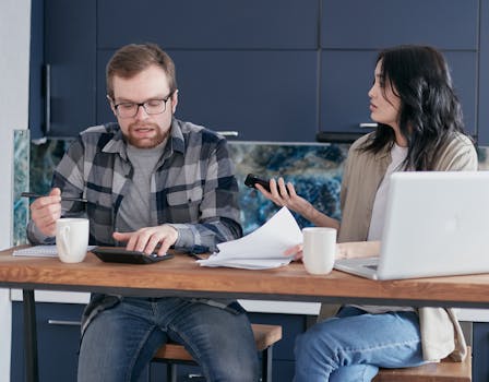 Two colleagues engaged in a work discussion around an office table, using a calculator and documents.
