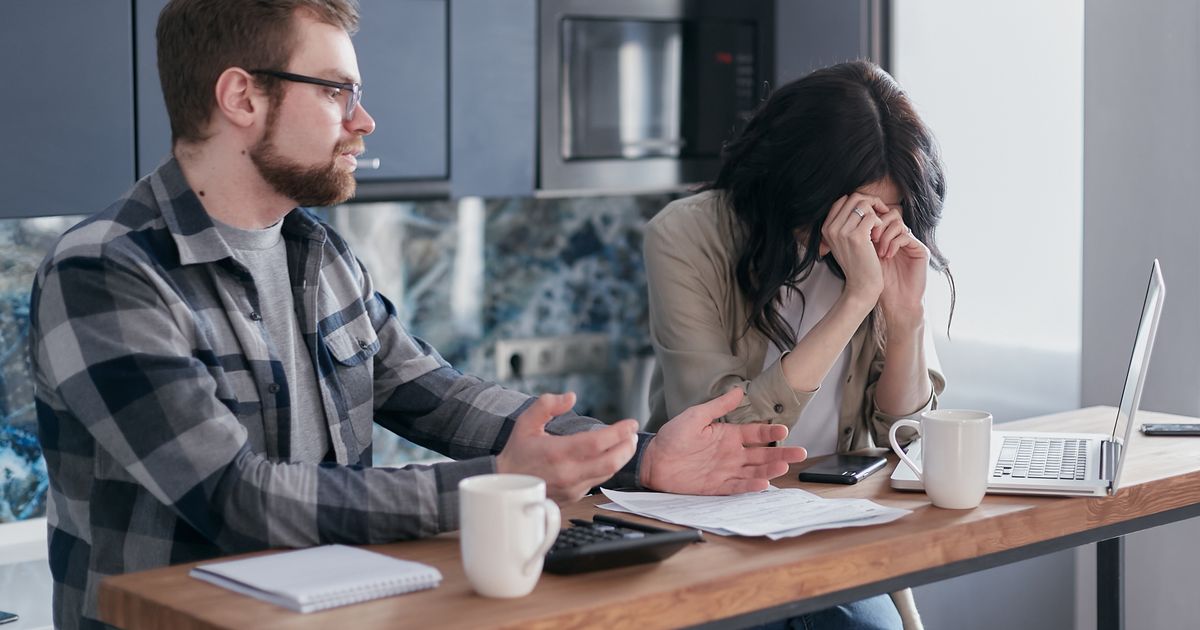 Person looking distressed at a kitchen table with a laptop and papers, being comforted