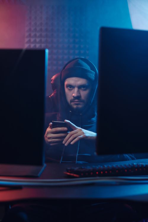 Free A mysterious hacker in a dark room using a smartphone between two monitors Stock Photo