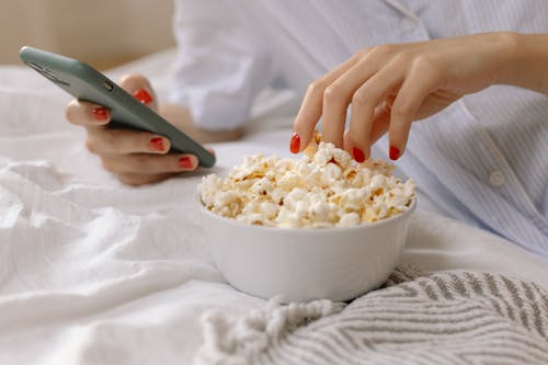 Person with Red Nails Using a Smartphone while Eating Popcorn