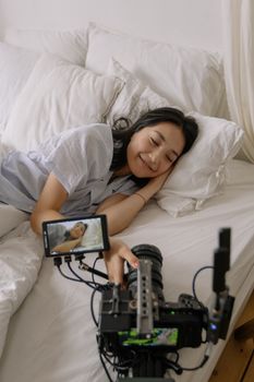 A woman lying in bed, taking a selfie with a camera and smiling in a cozy bedroom environment.