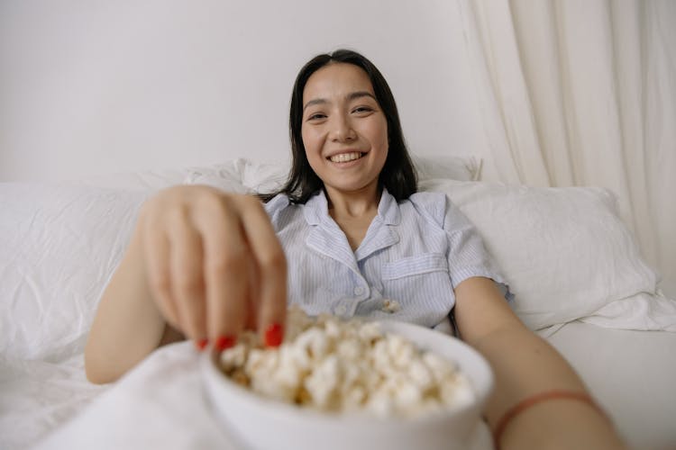 Woman In Pajamas Sitting On Bed Eating Popcorn